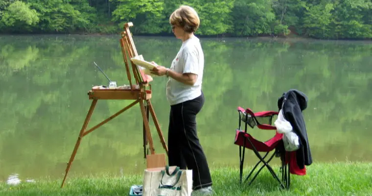 A woman from the Just Plein Nuts plein air painting group is painting on an easel near a lake.